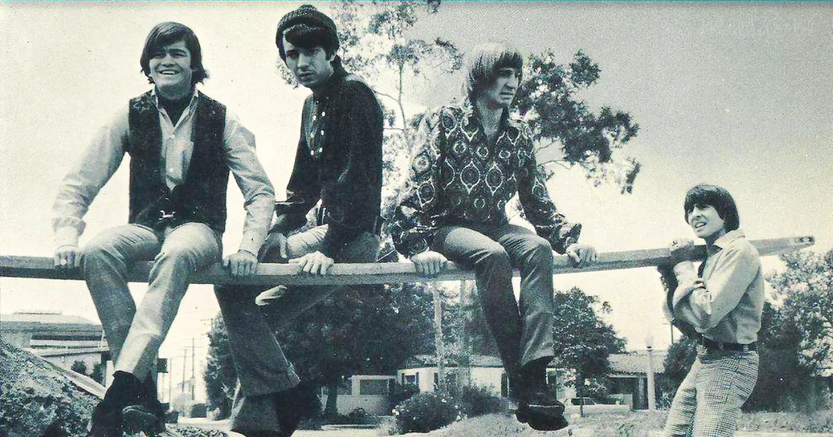 Four men in 1970s attire sit on a railing, smiling and posing outdoors.
