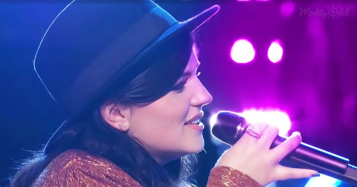 A person wearing a black hat and a sparkly top is singing into a microphone on stage with purple lighting.