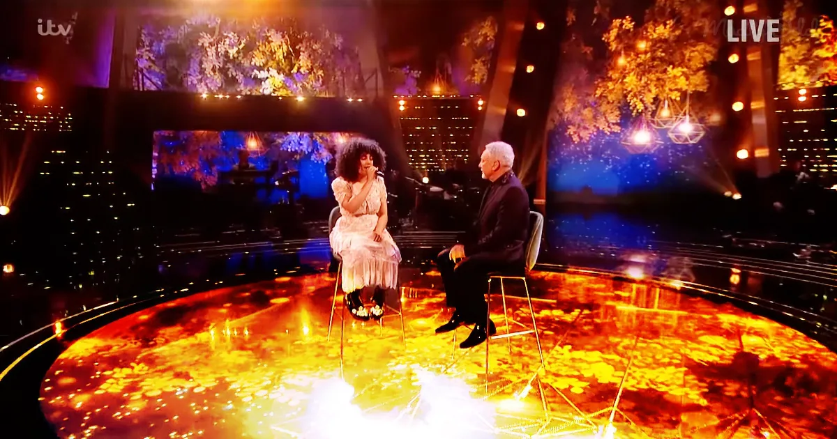 A woman in a white dress sits on a chair, talking to a man in a suit on a stage with a glowing floor and cityscape backdrop.