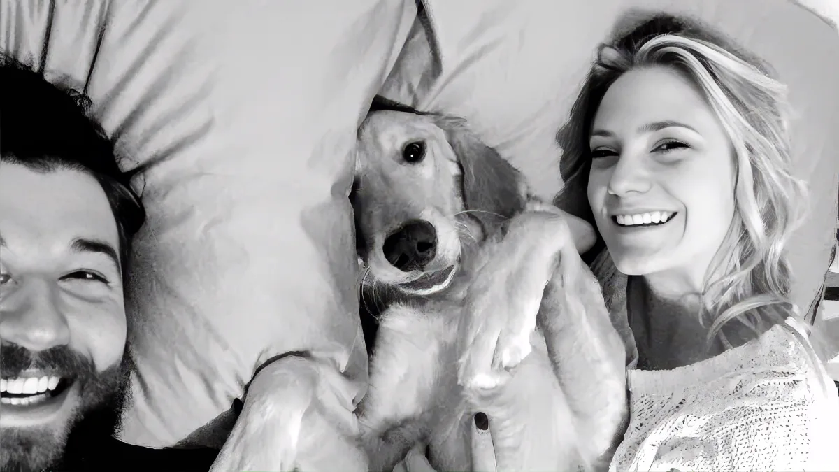 A couple and their dog are lying in bed, smiling and laughing together.