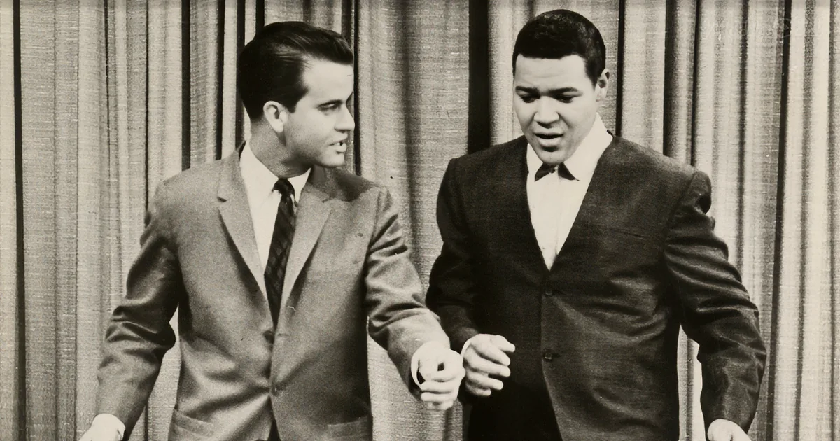 Two men in suits, one holding a microphone, standing in front of a curtain backdrop.