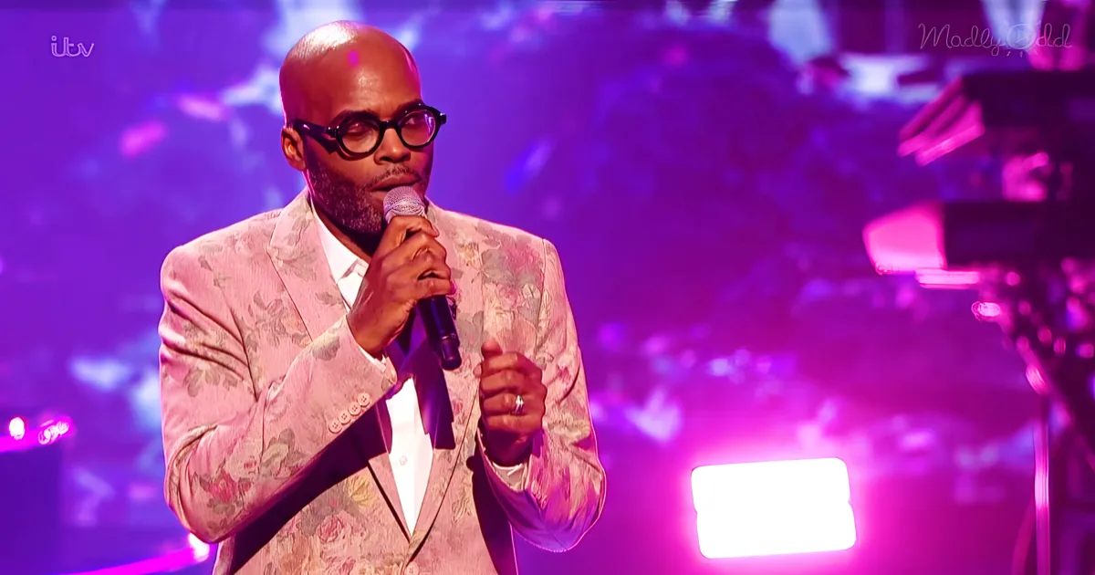 A man in a floral suit sings into a microphone on a stage with purple lighting.