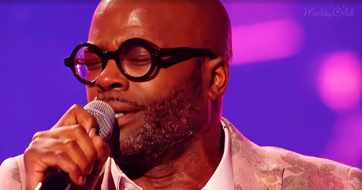 A man with glasses sings into a microphone, wearing a patterned jacket, against a purple-lit background.