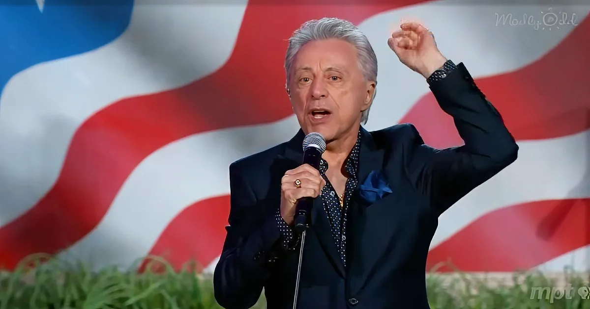 A man in a suit holds a microphone, raising his fist, with a red, white, and blue background.