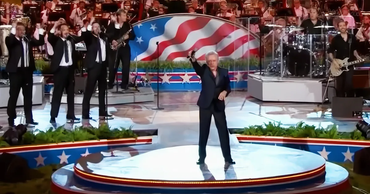 A man in a suit stands on a stage with a large American flag backdrop, surrounded by musicians.