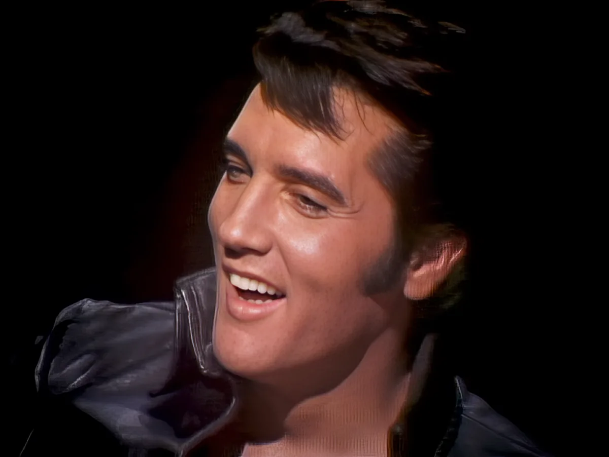 A smiling man with dark hair and a black leather jacket, set against a dark background.