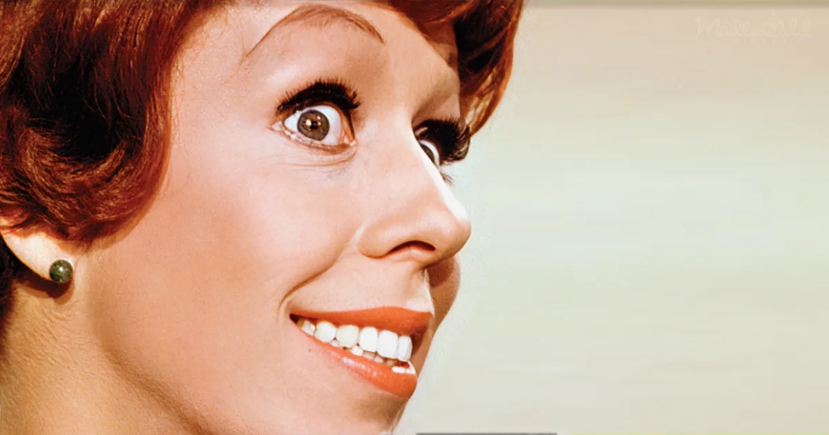 A smiling woman with red hair, wearing a green earring, and a light background.