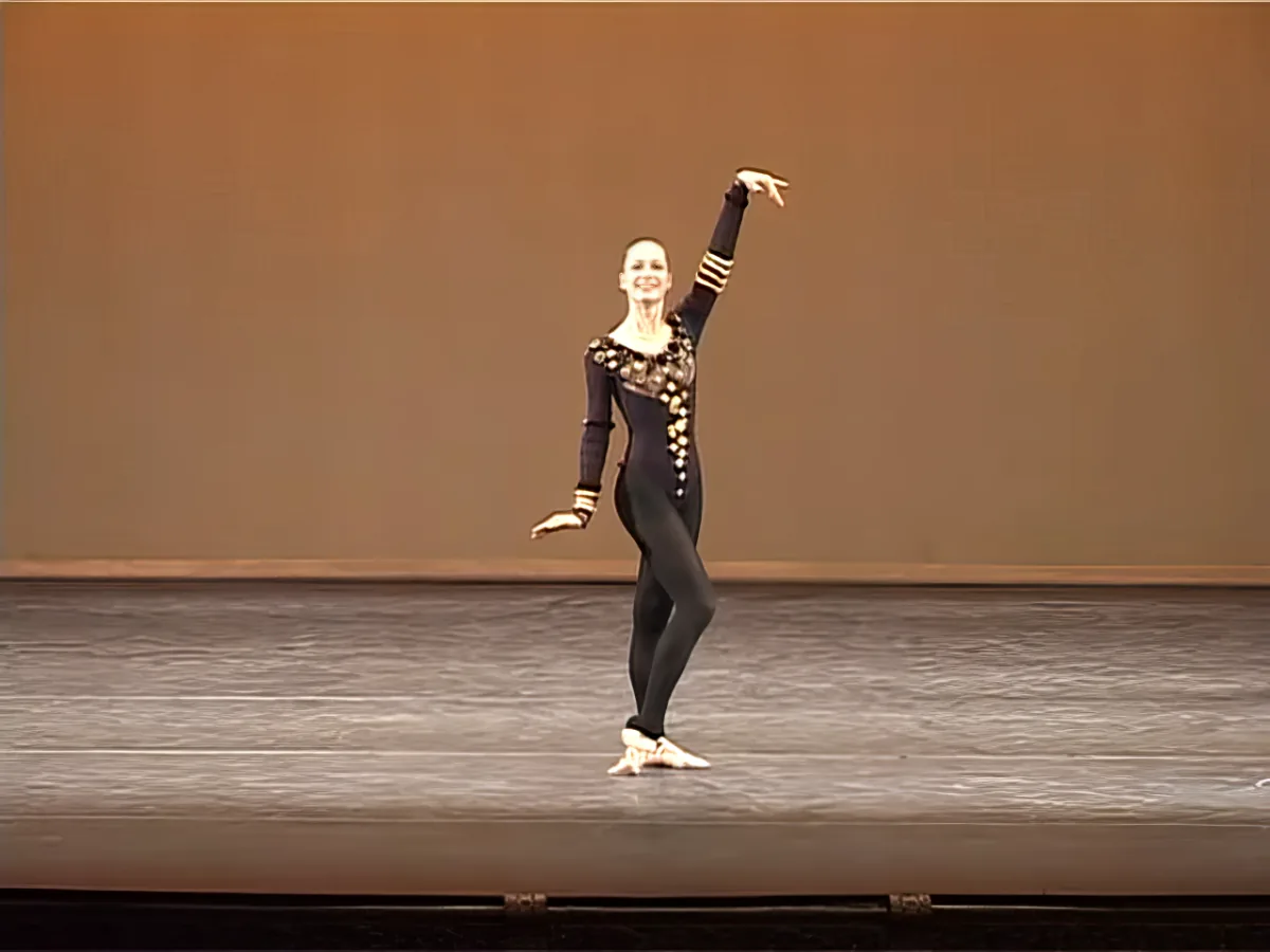A ballerina in a black leotard and tights performs a graceful pose on stage.