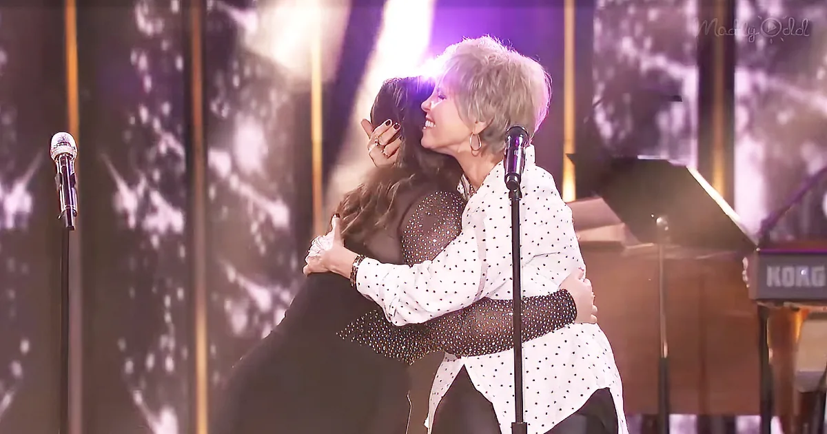 A woman in a polka-dot shirt hugs another woman on stage with microphones and a piano in the background.