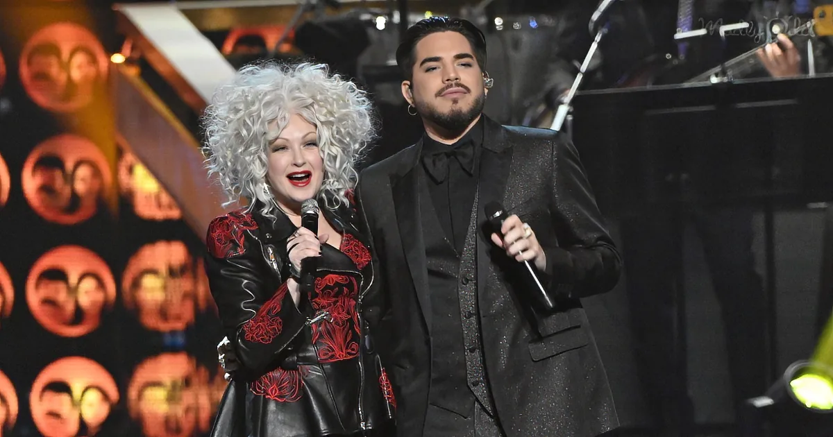 A woman with white curly hair and a man in a suit sing on stage, holding microphones.