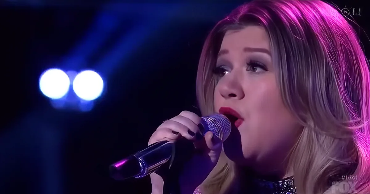 A person with long hair sings into a microphone under blue stage lights.