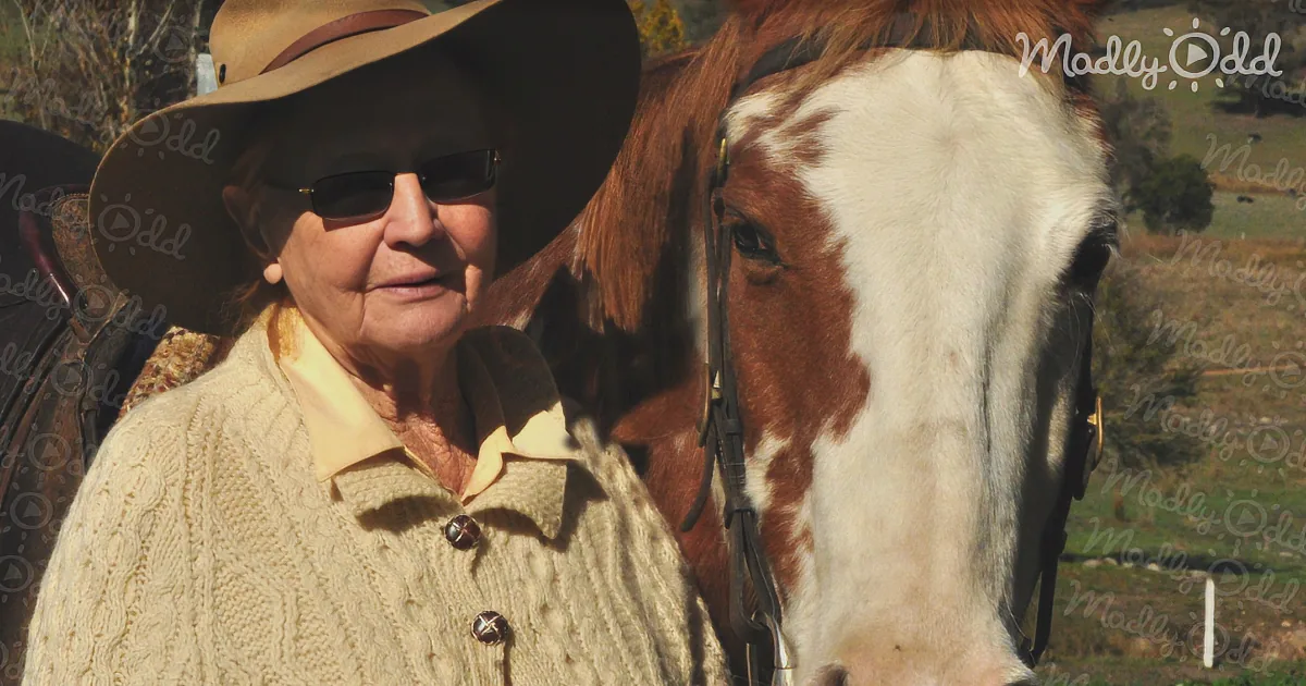 An elderly woman in a cowboy hat and sweater stands beside a horse with a white and brown coat.