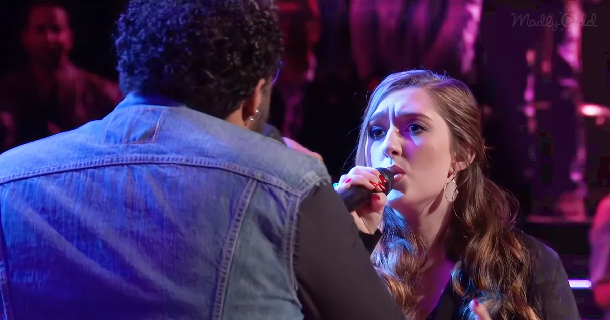 A woman sings into a microphone while a man in a denim jacket stands beside her.
