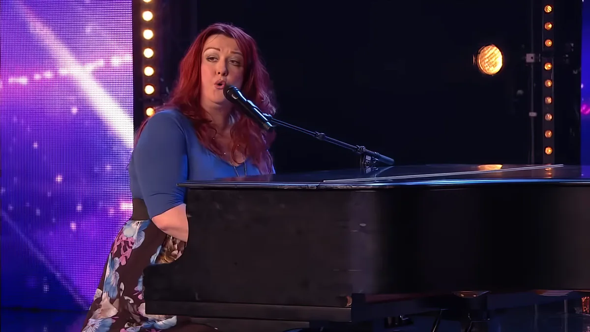 A woman with red hair sings into a microphone while playing a piano on a stage with purple lighting.