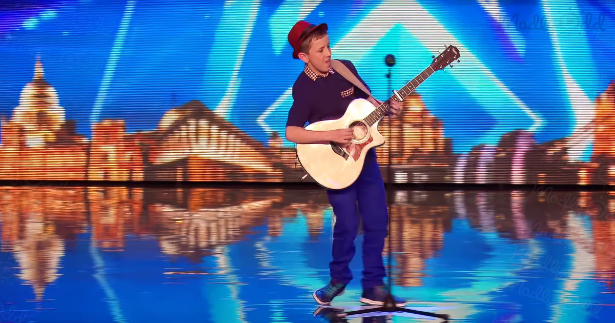 A young man in a red hat plays an acoustic guitar on a stage with a cityscape backdrop.