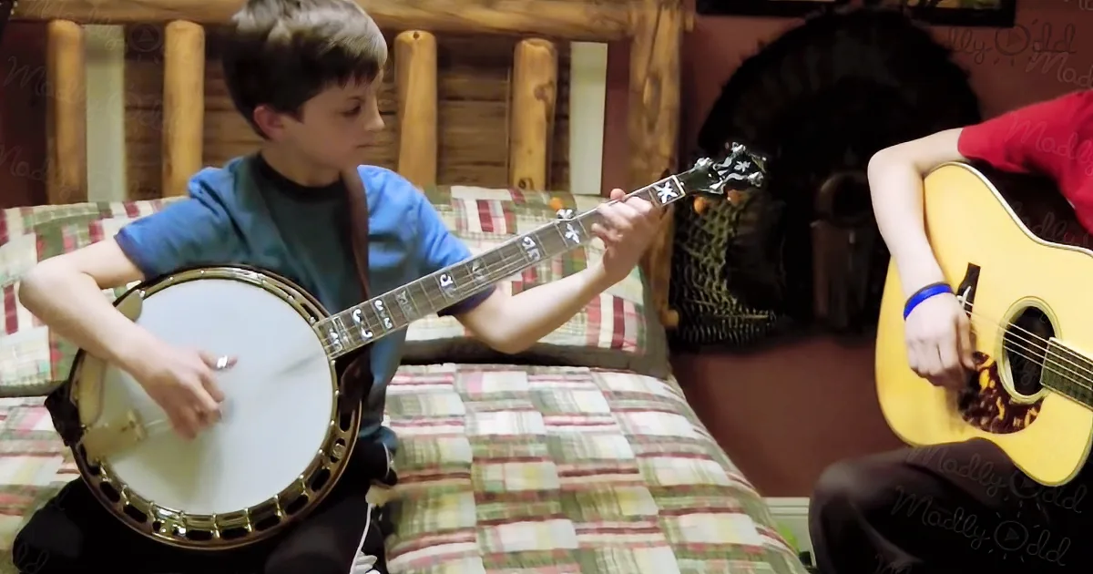 A boy plays a banjo while another plays an acoustic guitar in a cozy room.