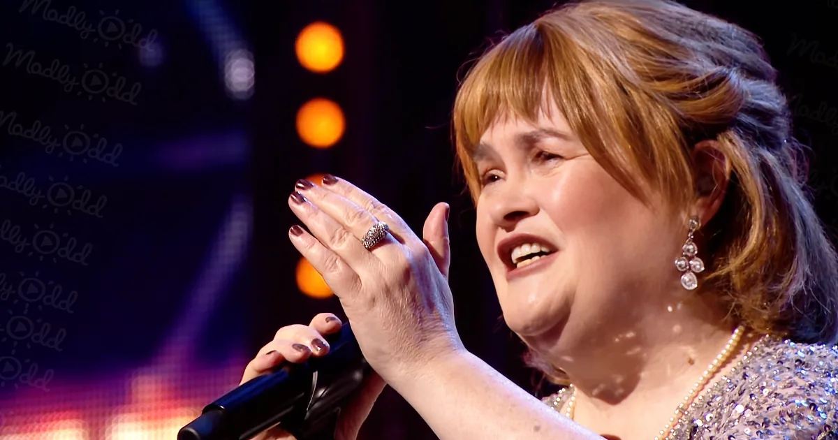 A woman with short brown hair, wearing a pearl necklace and earrings, sings into a microphone on stage.