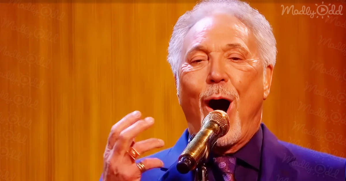 An older man with gray hair and a beard sings into a microphone, wearing a blue shirt and rings.