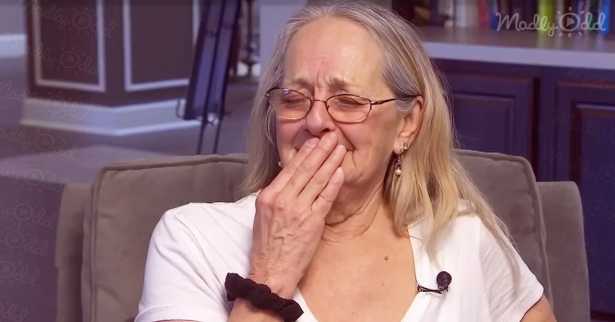 An older woman with long blonde hair, wearing glasses and a white shirt, sits on a couch, covering her mouth with her hand.