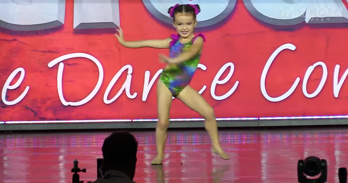 A young girl in a colorful leotard dances gracefully on stage, smiling.