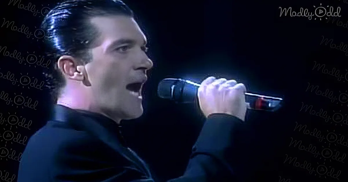 A man in a black shirt sings passionately into a microphone, illuminated by stage lighting.