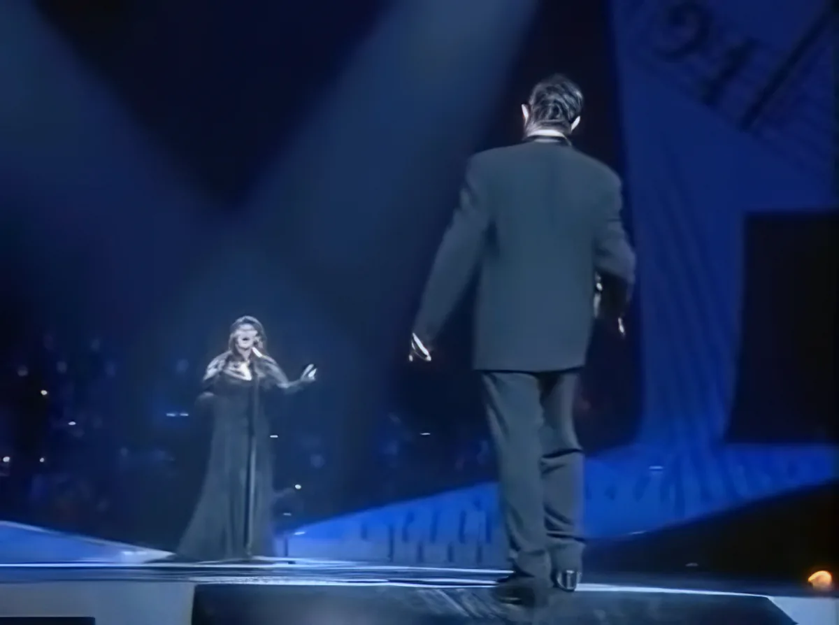 A man in a suit walks across a stage, facing a woman singing into a microphone.
