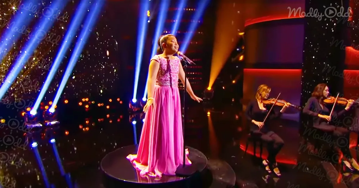 A young girl in a pink dress sings on stage with a microphone, accompanied by musicians playing violins.