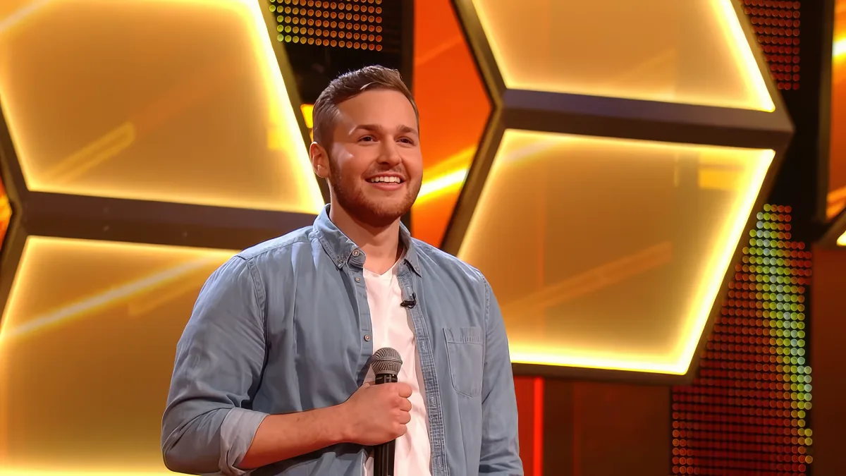 A man in a blue shirt holds a microphone, smiling, with a colorful, geometric background.