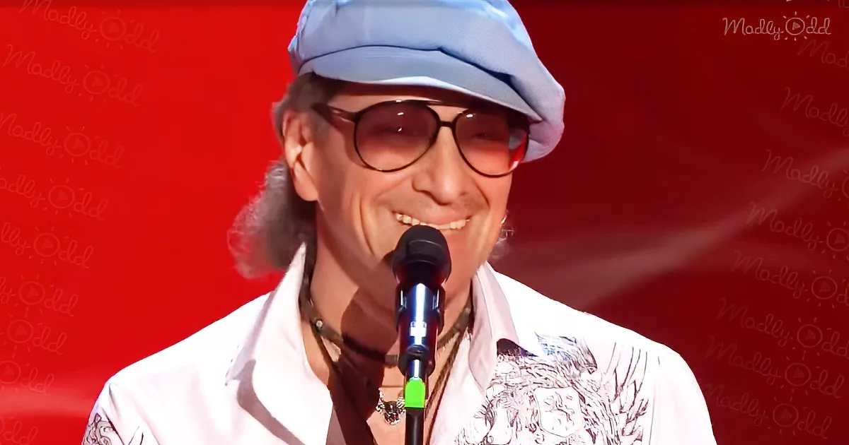 A smiling person wearing a white shirt, cap, and sunglasses stands in front of a microphone against a red background.