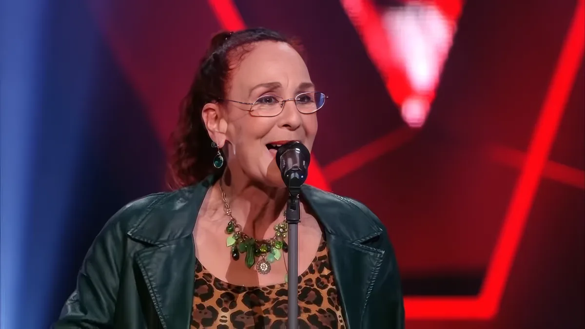 A woman in a leopard print top and green jacket sings into a microphone on stage with red and blue lighting.