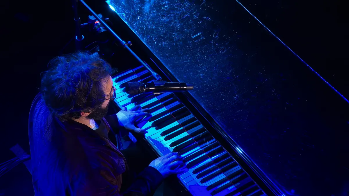 A musician plays a grand piano under blue stage lighting, with a microphone positioned above the keys.