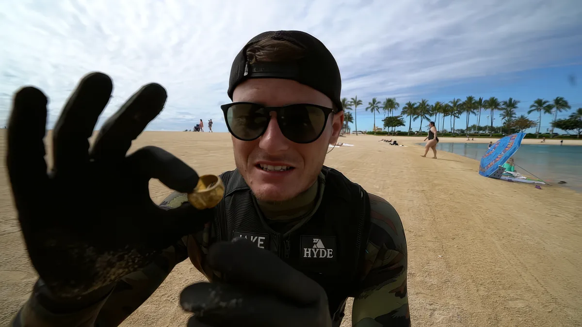 A person wearing a black cap, sunglasses, and a camouflage wetsuit holds up a small object on a sandy beach with palm...