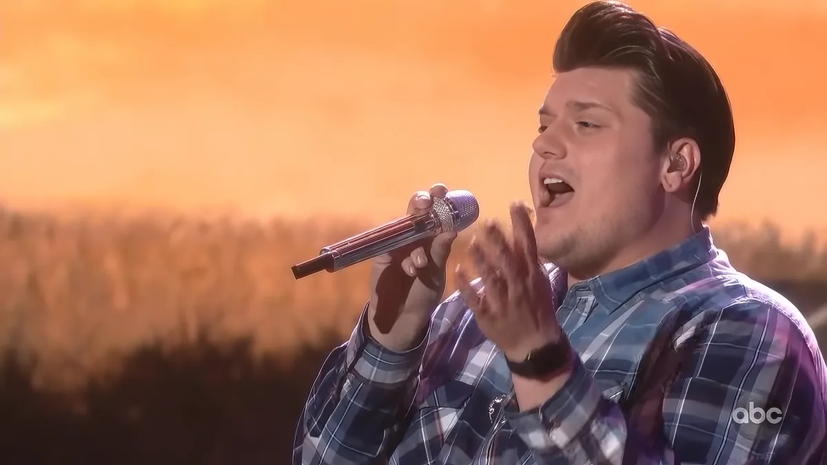 A man in a plaid shirt sings passionately into a microphone against a blurred field backdrop.