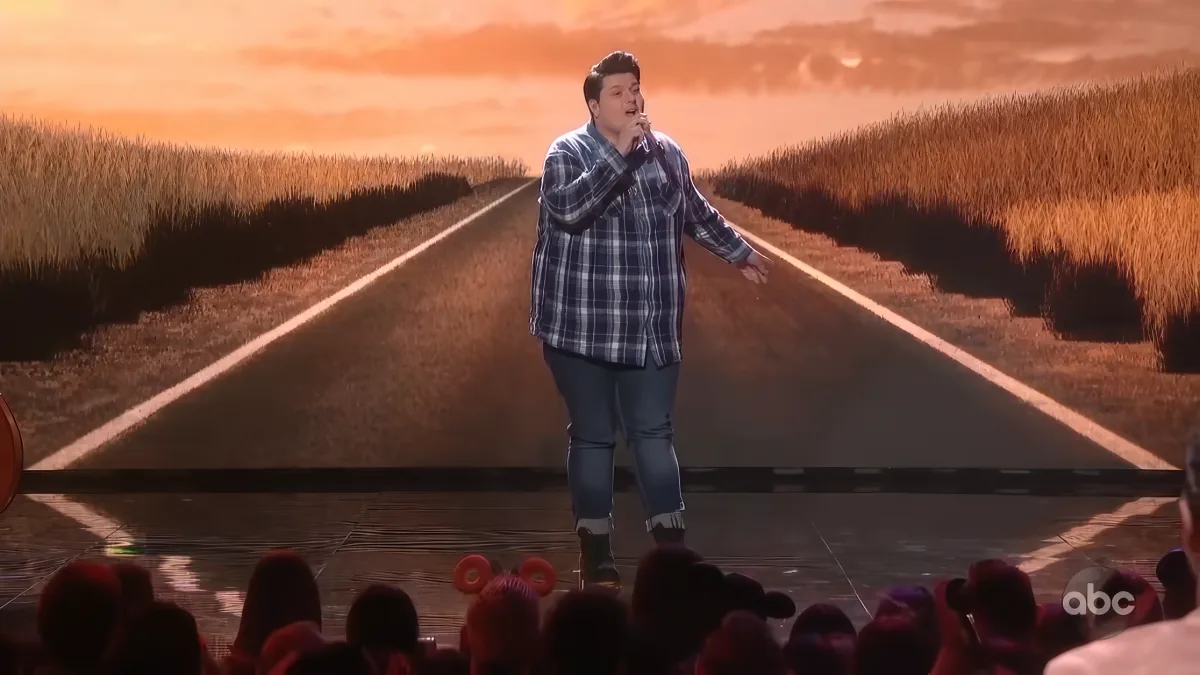 A man in a plaid shirt sings on stage with a backdrop of a road and wheat fields.