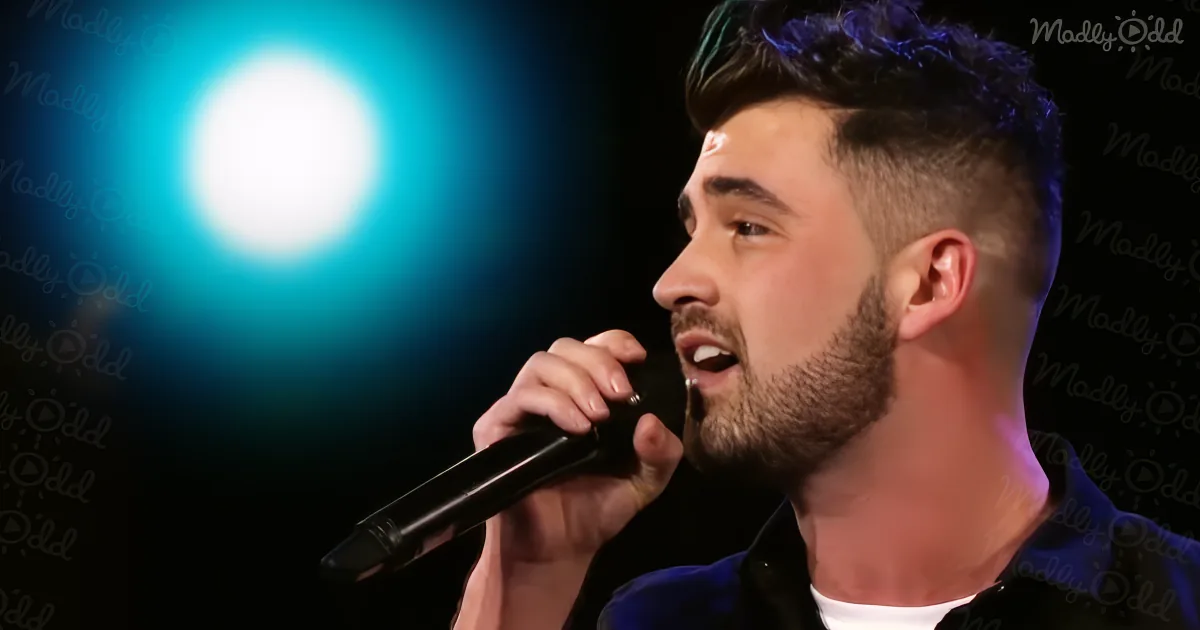 A man with short hair and a beard sings into a microphone under a bright blue spotlight.