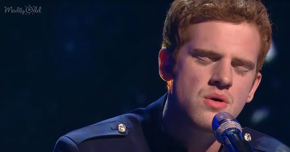 A man with short red hair and a blue jacket sings into a microphone with his eyes closed.