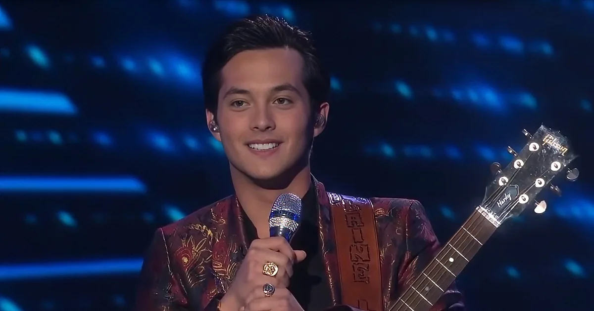 A man in a patterned jacket holds a microphone and a guitar, smiling on stage with blue lights.