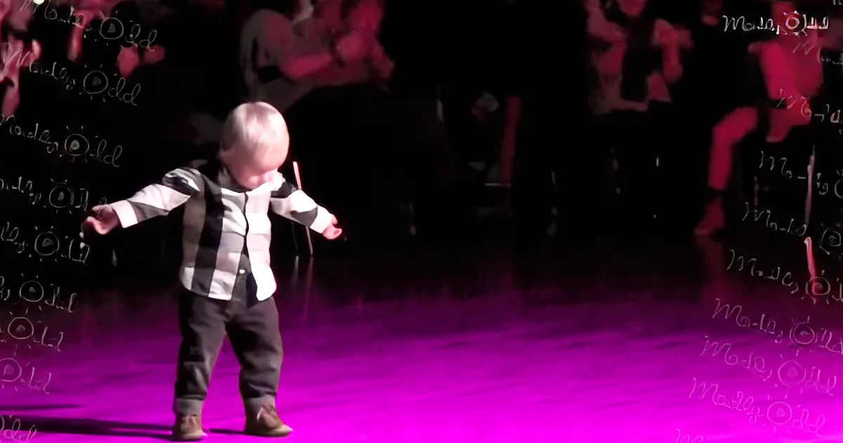 A child in a striped shirt and pants stands on a purple-lit stage with arms outstretched.