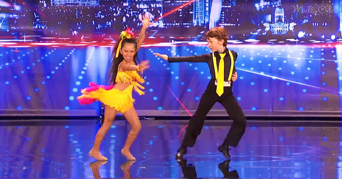 A young girl in a yellow dress dances with a boy in a black suit on a stage with a cityscape backdrop.