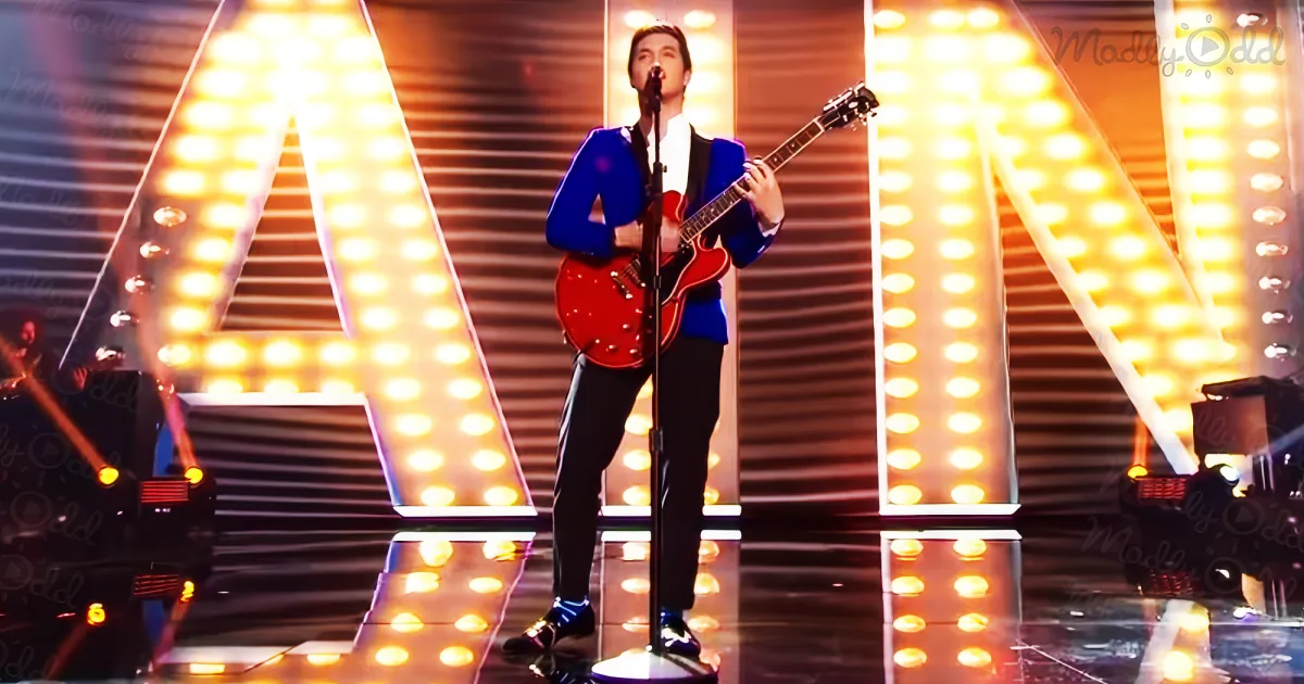 A man in a blue suit plays a red guitar on stage with bright yellow letter lights behind him.