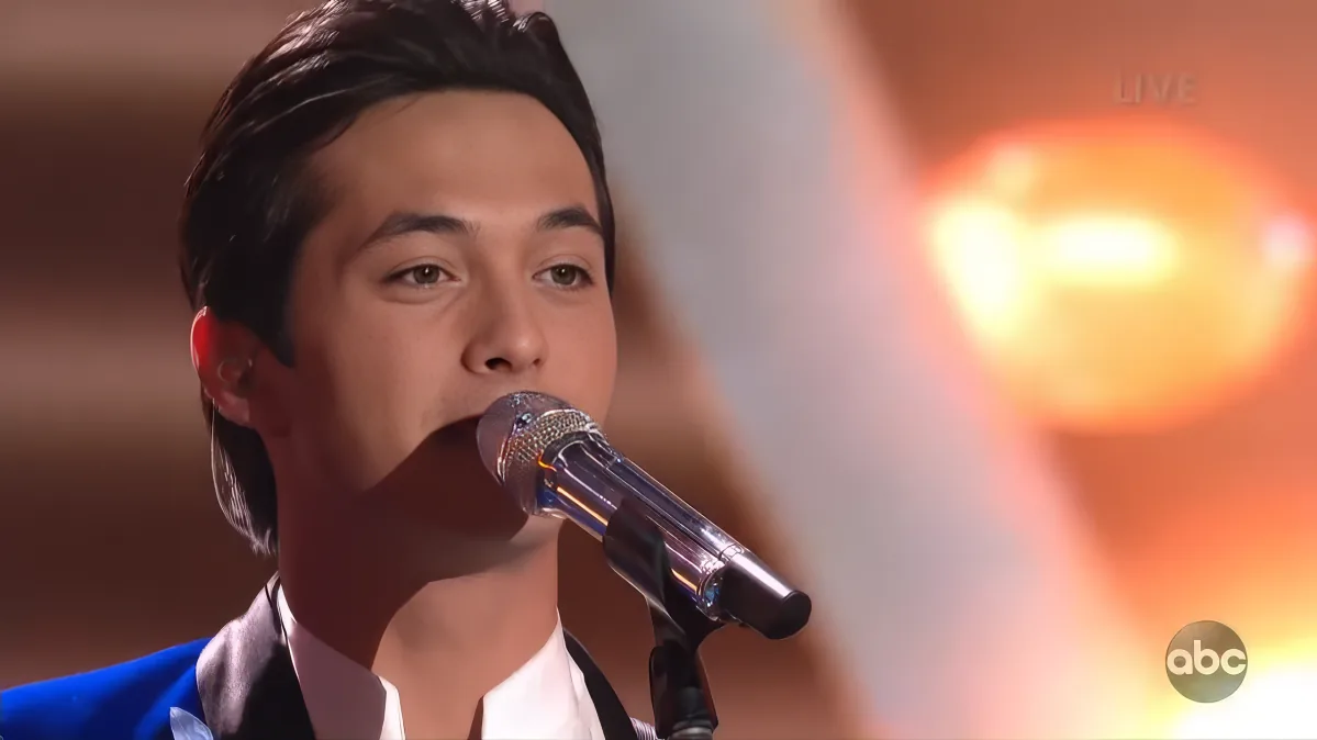A man in a suit sings into a microphone, with a bright light and "LIVE" text in the background.