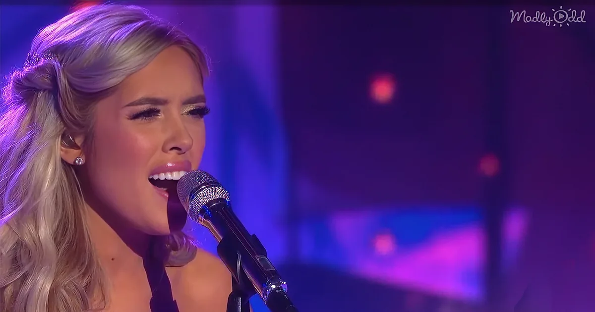 A woman with long blonde hair sings into a microphone on stage with purple lighting.