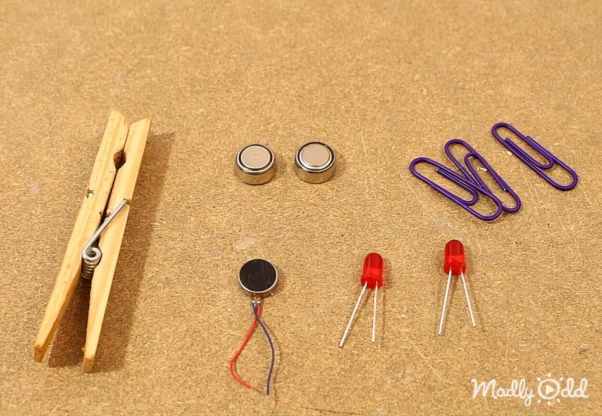 A clothespin, two small batteries, a paperclip, and two red LEDs on a brown surface.