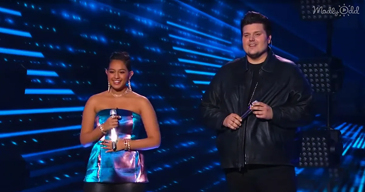 A woman in a shiny dress and a man in a black jacket stand on stage holding microphones, smiling.