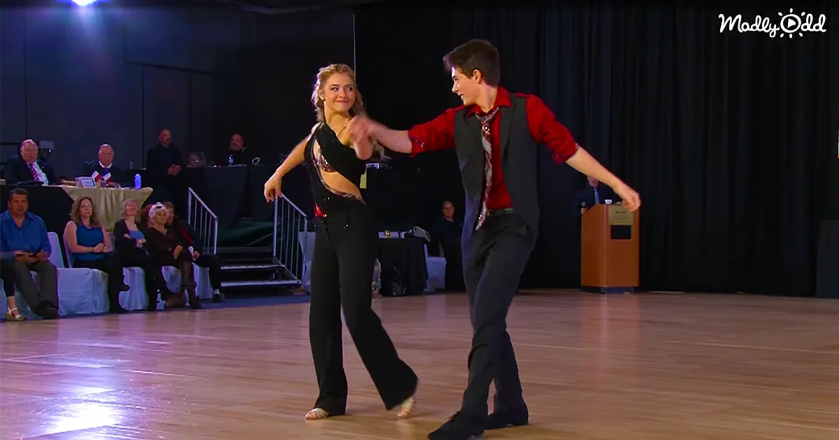 A couple in formal attire dances gracefully on a wooden floor, surrounded by spectators.