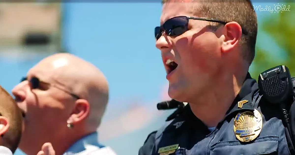 A police officer in uniform speaks into a microphone, wearing sunglasses and a badge, with another person in the background.