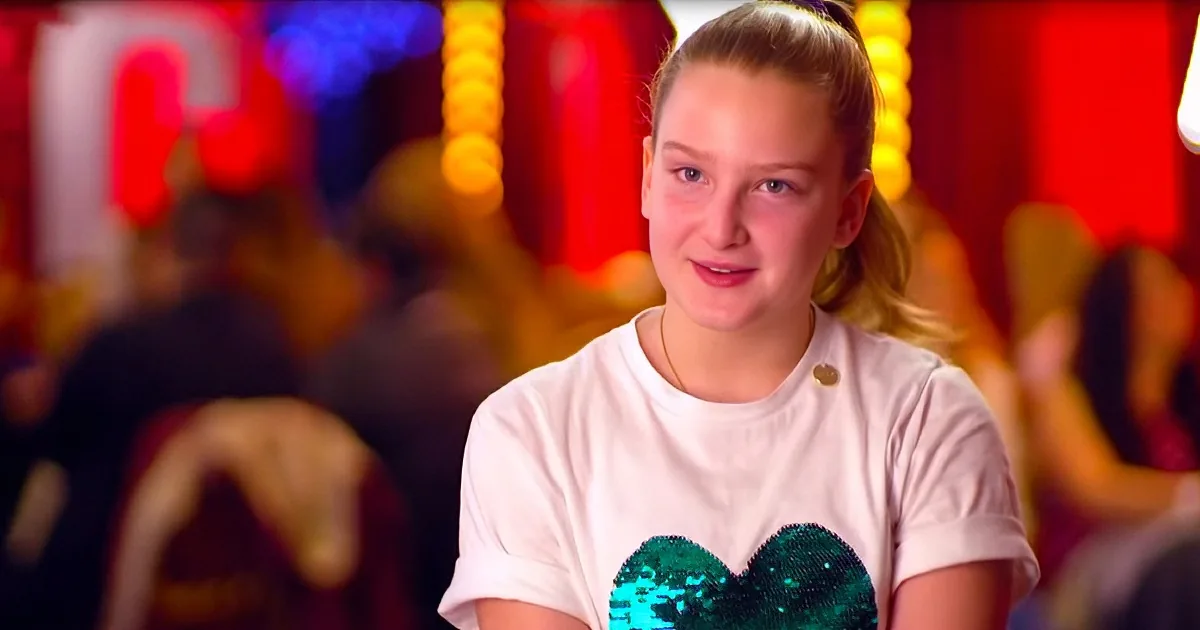A person in a white t-shirt with a green sequined heart stands in front of a colorful, blurred background.