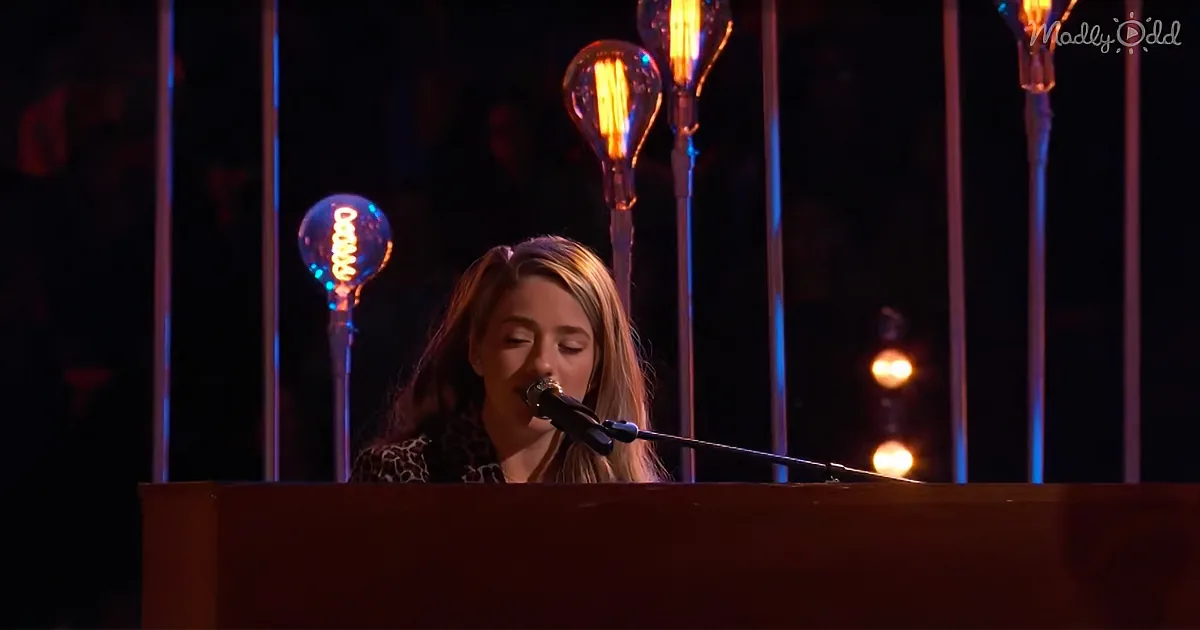 A person singing into a microphone at a piano, surrounded by illuminated light bulbs.