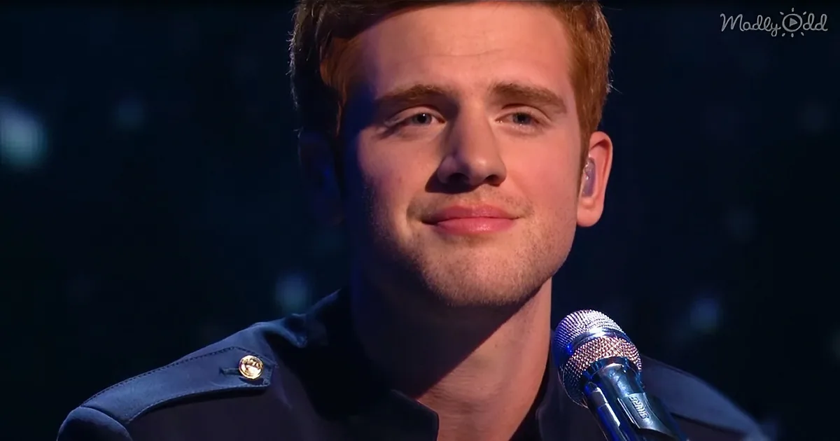 A man with short brown hair and a beard, wearing a dark jacket, is singing into a microphone.