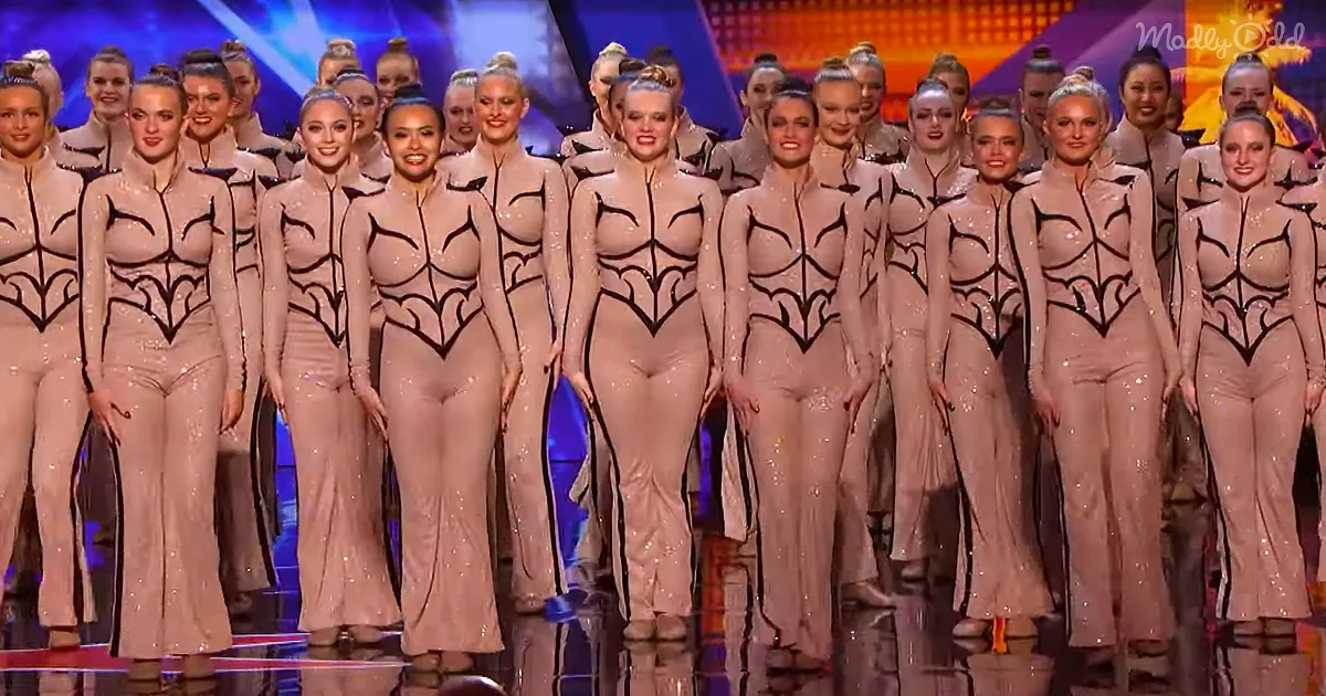 A group of performers in matching beige outfits with black designs stand in formation on a stage.